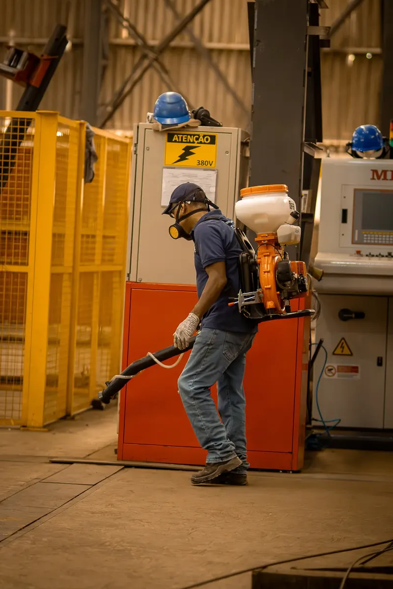Profissional com uniforme azul-marinho e equipamento costal próximo a painéis elétricos em um galpão.