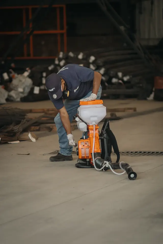 Profissional com uniforme azul-marinho e máscara preparando um equipamento costal no piso de um galpão.