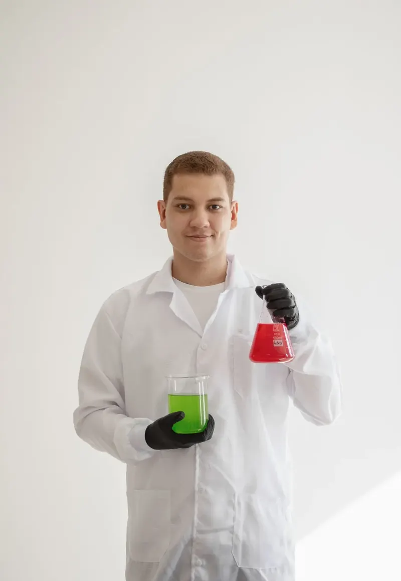 Klauver Yuri S. Melo, de jaleco branco, segurando um béquer com líquido verde e um frasco com líquido vermelho.