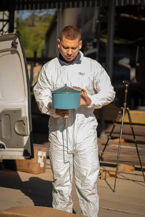 Profissional de macacão branco segurando um recipiente azul com tampa, ao lado do veículo da empresa.