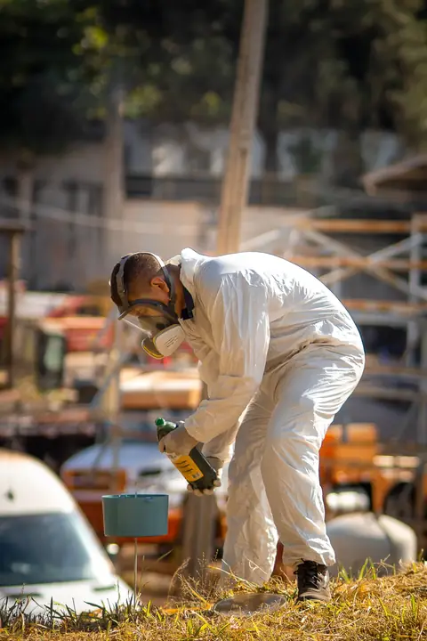Profissional de macacão e máscara de proteção segurando um frasco junto a um recipiente instalado em área externa.