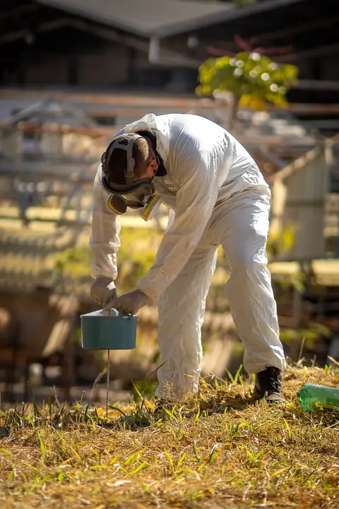 Profissional de macacão e máscara de proteção inclinado sobre um recipiente instalado no gramado.