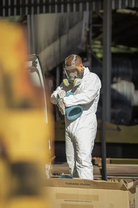 Profissional de macacão e máscara de proteção segurando a tampa de um recipiente em área coberta, junto a caixas.