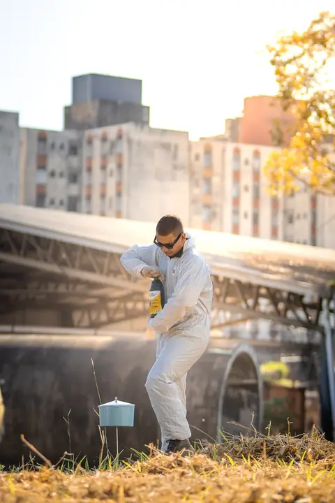 Profissional de macacão e óculos escuros em área externa, junto a um recipiente instalado, com prédios ao fundo.