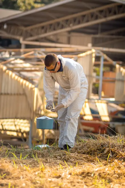 Profissional de macacão, óculos e luvas manuseando um recipiente instalado sobre vegetação seca.