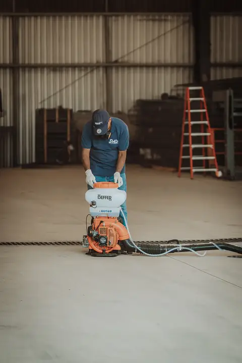 Profissional com uniforme azul ajustando um equipamento motorizado no piso de um galpão.