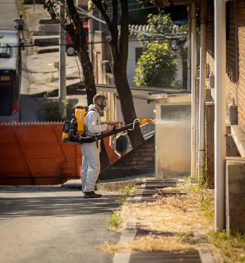Profissional de macacão operando um equipamento costal em área externa, junto a um muro e uma calçada.