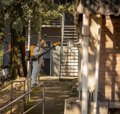 Profissional de macacão com equipamento costal em uma passagem externa com escada e corrimão.