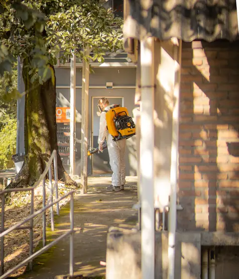 Profissional de macacão com equipamento costal em um corredor externo com corrimão e árvores.