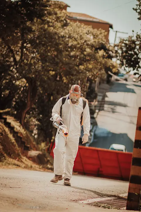 Profissional de macacão e máscara facial caminhando com um equipamento portátil em área externa.