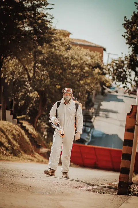 Profissional de macacão e máscara facial caminhando por uma via externa com um equipamento portátil.