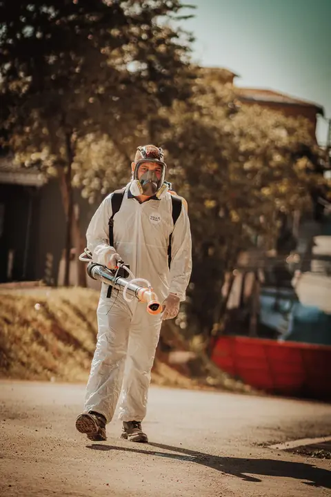 Profissional de macacão e máscara facial segurando um equipamento portátil em área externa arborizada.