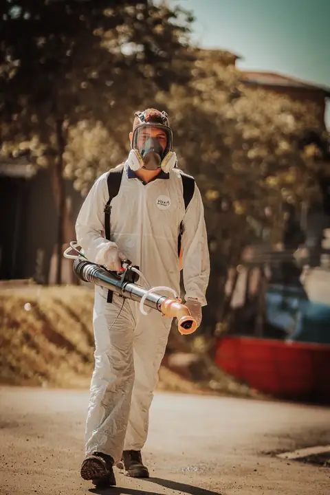 Profissional de macacão e máscara facial caminhando em direção à câmera com um equipamento portátil.