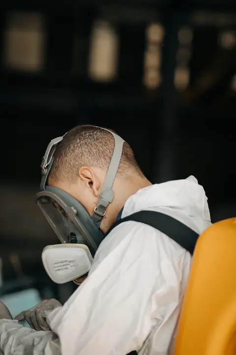 Profissional de macacão ajustando a máscara de proteção respiratória, com equipamento costal amarelo.