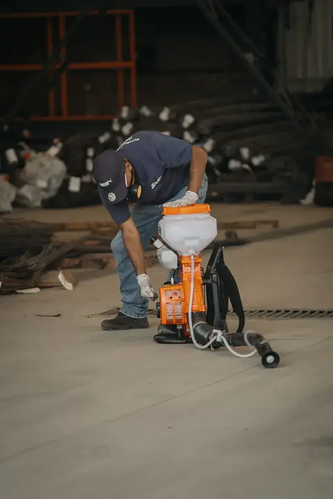 Profissional com uniforme azul-marinho e máscara preparando um equipamento costal no piso de um galpão.