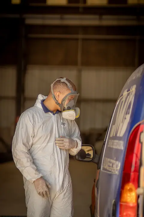 Profissional de macacão e máscara facial ao lado do veículo da empresa, dentro de um galpão.