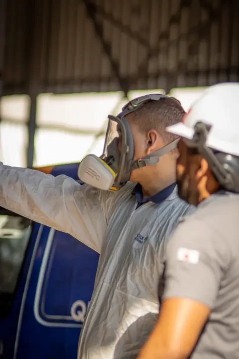 Profissional de macacão e máscara facial conversando com uma pessoa de capacete branco e apontando para o ambiente.
