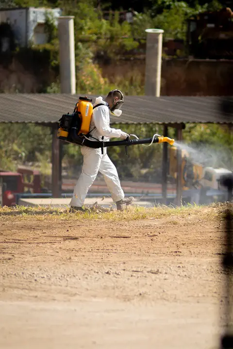 Profissional de macacão operando um equipamento costal em área externa, com névoa visível.