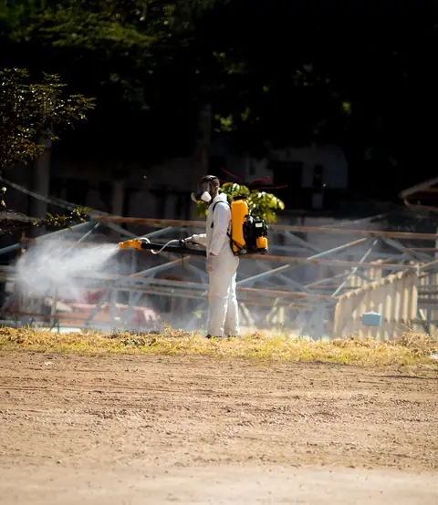 Profissional de macacão operando um equipamento costal em área externa aberta, com névoa visível.