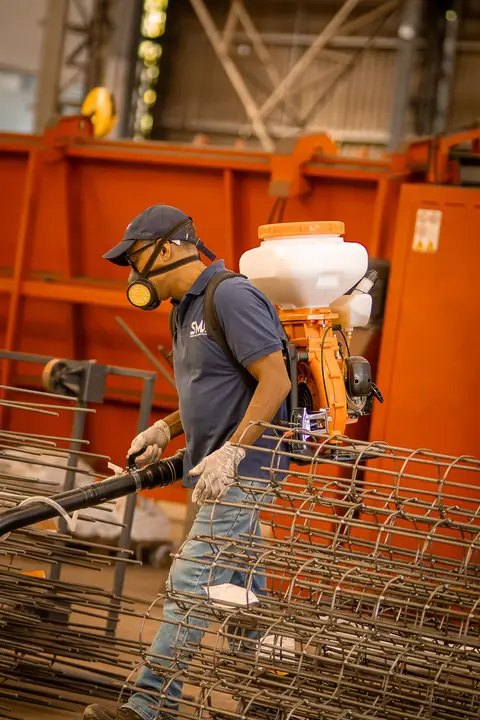 Profissional com uniforme azul-marinho, máscara e equipamento costal entre estruturas de vergalhões em um galpão.