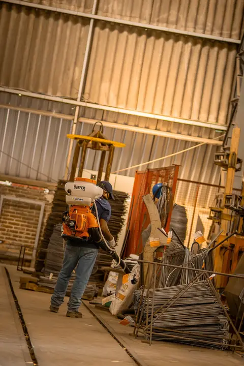 Profissional com equipamento costal em um galpão com materiais armazenados.