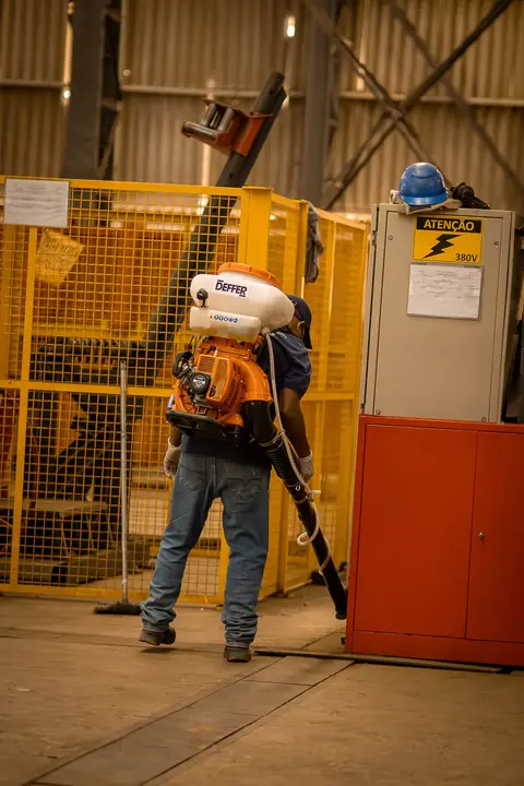 Profissional de costas, com equipamento costal, próximo a grades amarelas e painéis elétricos em um galpão.