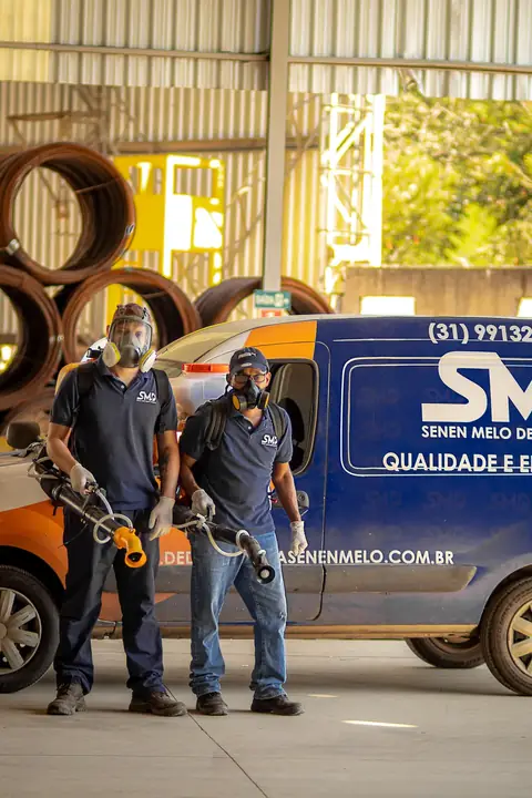 Dois profissionais com máscaras faciais e equipamentos portáteis em frente ao veículo da Senen Melo, em um galpão.